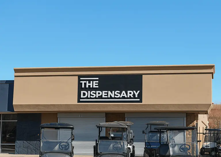 Exterior of "The Dispensary" storefront on a clear day, with beige stucco walls, black-and-white signage, with golf carts in front.